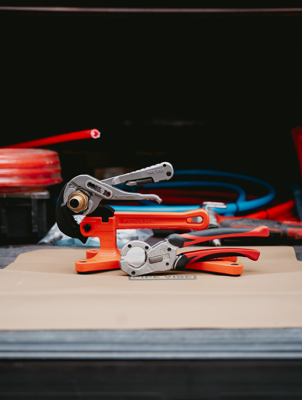 A set of plumbing tools including an orange and silver pipe crimping tool and a red and black pipe cutter, displayed on a cardboard surface with a dark, blurred background.